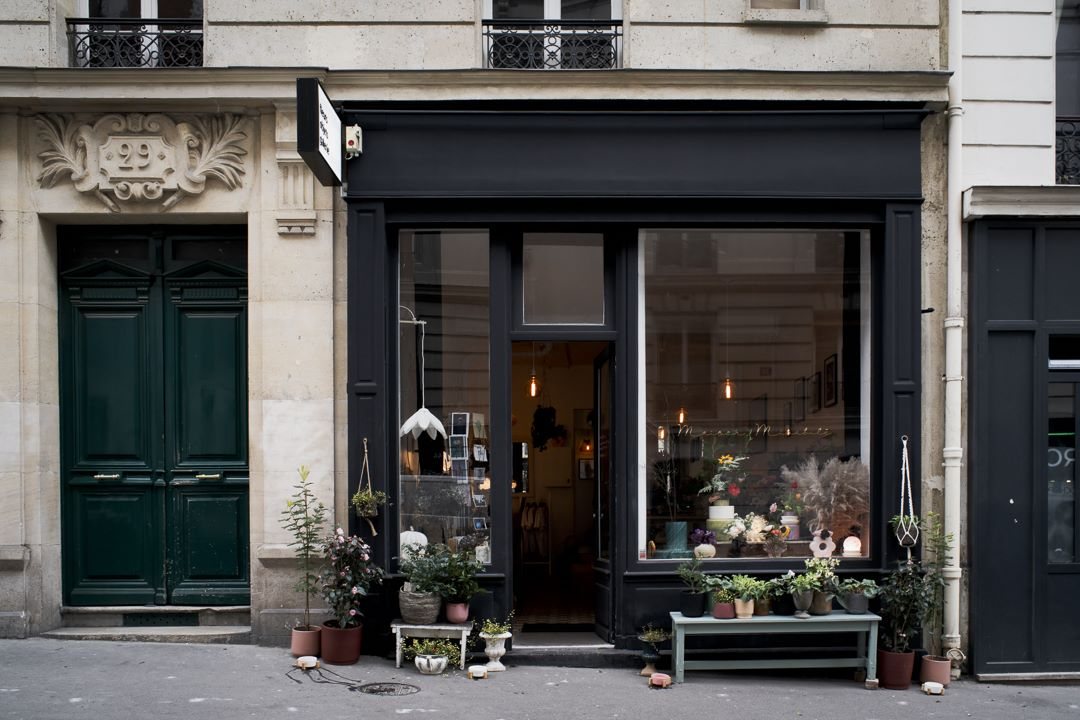 La boutique vue de la rue, façade noire entre la porte cochère et les pots posés sur le trottoir