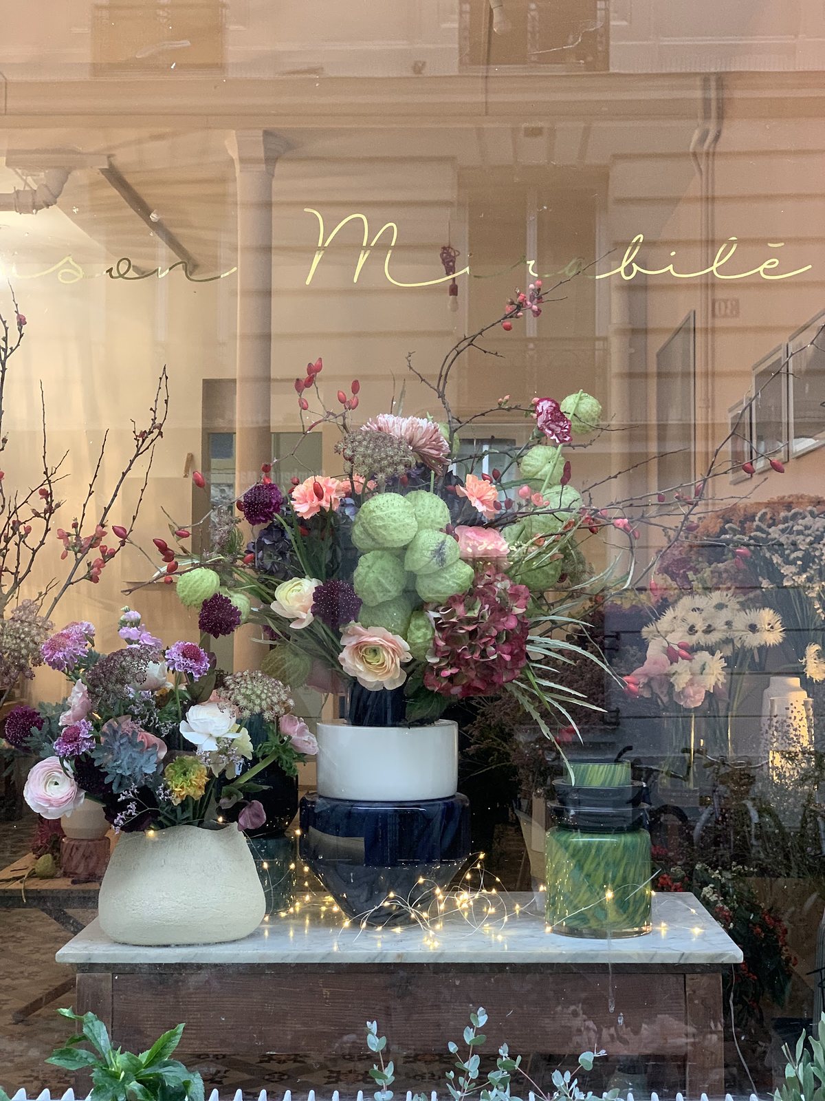 Vitrine de printemps : bouquets de renoncules et de dahlias posés devant une guirlande lumineuse